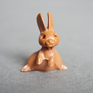 May include: A small, brown plastic rabbit figurine with red eyes. The rabbit is sitting upright with its ears perked up.