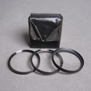 May include: A set of three clear camera lens filters with black metal rims, arranged in front of a black leather case with a button closure. The filters are circular and appear to be unused, with a gray background.