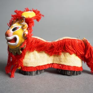May include: A decorative Chinese lion dance figure. The lion has a yellow face with red and white accents, a red mane, and a cream-colored body. The lion's feet are black and the figure is on a gray background.