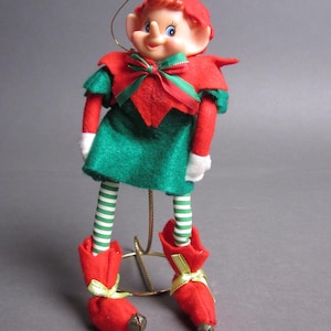 May include: A red and green Christmas elf ornament with a green and white striped stocking and red boots with bells. The elf has a red hat, a green and red bow, and a white face with blue eyes.
