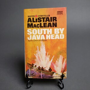 May include: A vintage paperback book titled "South By Java Head" by Alistair MacLean. The cover features a boat with figures, set against an orange and yellow background with white bursts. The book is displayed on a black stand.