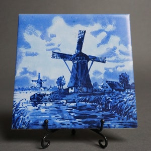 May include: A blue and white ceramic tile depicting a Dutch windmill scene. The tile features a large windmill with two smaller windmills in the background, all set against a blue sky with white clouds. The scene also includes a river, trees, and grass.