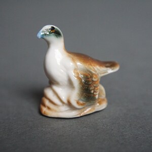 May include: A small ceramic figurine of a brown and white bird with a blue eye. The bird is perched on a small base.