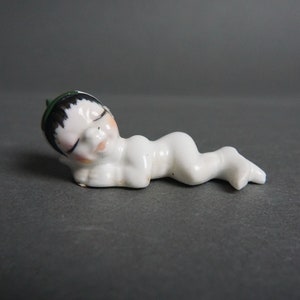 May include: A small white porcelain figurine of a sleeping baby wearing a green hat. The baby is lying on its side with its arms crossed and its eyes closed.