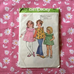 May include: A vintage Simplicity sewing pattern for a child's dress, size 2. The pattern envelope shows illustrations of three young girls in various dresses. The background is a pink and white floral pattern.