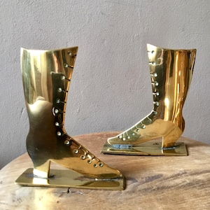 May include: Two vintage brass boot sculptures. Each boot is a shiny gold colour, with a high-top design and a lace-up front. The boots are mounted on rectangular bases and sit on a wooden surface.