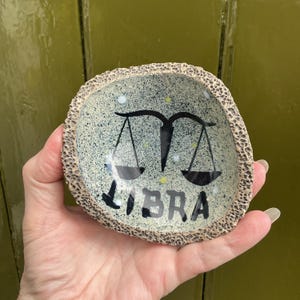May include: A small, speckled ceramic bowl with a textured rim. The bowl features a black Libra symbol and the word "LIBRA" in black lettering. The interior has a light blue and white speckled design.