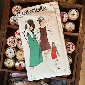 May include: Vintage sewing pattern for a pinafore dress and blouse by Maudella. The pattern features illustrations of the dress in green and red, with a white blouse. The pattern is labeled "5520" and includes measurements. The image is surrounded by spools of thread.