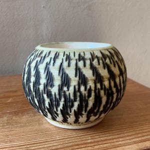 May include: A white ceramic mug with a black handle and the text "But First, Coffee" in a bold, black font. The mug is set against a blurred background, suggesting a cosy setting.