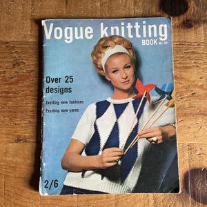 以下が含まれることがあります： ヴィンテージヴォーグ編み物ブックNo. 62。表紙には、白とネイビーブルーのアーガイルセーターを着た女性がいます。「Over 25 designs」と「Exciting new fashions」と書かれています。本は木製の表面にあります。