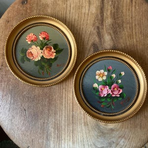 May include: Two circular wall art pieces featuring hand-painted floral arrangements. Each piece is encased in a gold-toned frame with a beaded inner edge. The left painting displays peach and coral roses, while the right shows pink and white flowers.