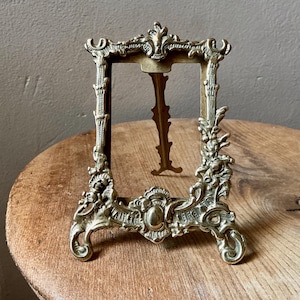 May include: A rectangular brass picture frame featuring an ornate, embossed metal border with intricate scrollwork and floral motifs. The frame includes a central vertical opening and a hinged metal stand attached to the back for upright display.
