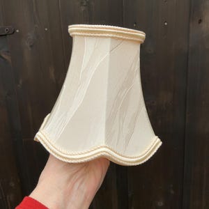 May include: Cream-colored lampshade with a textured pattern and a scalloped edge. The lampshade has a beige trim around the top and bottom. The lampshade is held in a hand.