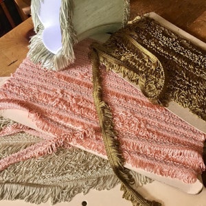 May include: An assortment of decorative fringe trim in various colors. The image shows rolls of pink, gold, and light green fringe trim. The trim has a textured appearance with a fringe edge, suitable for crafts and sewing projects.