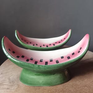 Peut inclure: Deux bols de service en céramique en forme de pastèque. Les bols ont un intérieur rose avec des graines carrées noires et un extérieur vert. Les bols sont sur un socle vert et sont placés sur une surface en bois.