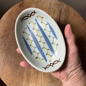 May include: Oval ceramic dish with a white base, featuring a geometric pattern of blue, yellow, and brown accents. The dish has a slightly raised rim and is held by a hand. The design includes diagonal blue stripes and zig-zag yellow lines.