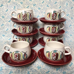 May include: A set of eight ceramic coffee cups and saucers. The cups are off-white with a colourful pineapple design. The saucers are a deep red. The cups have a rounded shape with a handle.