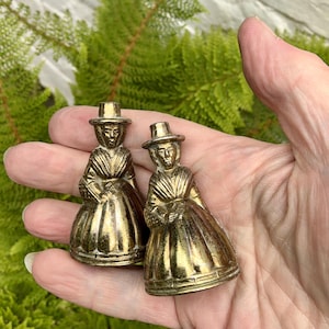 May include: Two small, metallic gold-toned figurines shaped like figures wearing wide-brimmed hats and long, pleated dresses. The figures have defined facial features and textured clothing details, with a polished finish that reflects light.