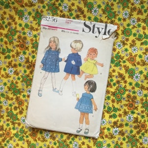 May include: Vintage toddler dress sewing pattern, size 1, with illustrations of children's dresses. The pattern envelope is white with the word "Style" in a large black font. The background is a yellow floral fabric.