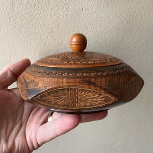 Könnte beinhalten: Ein runder Holzbehälter mit Deckel, der mit aufwendigen, handgeschnitzten geometrischen Mustern und Sonnenstrahlmotiven verziert ist. Das dunkelbraune Holz hat eine polierte Oberfläche und der Deckel besitzt einen kleinen, glatten Kugelknauf.