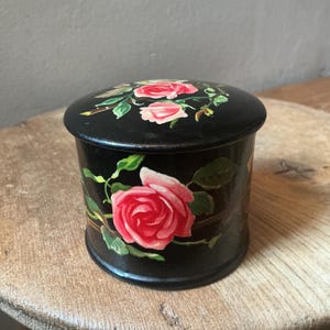 May include: A black, round trinket box with a lid, adorned with hand-painted pink roses and green leaves. The floral design is set against a black background, creating a vintage aesthetic. The box is sitting on a wooden surface.