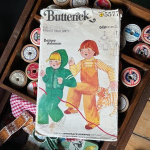 May include: Vintage Butterick sewing pattern, size 3, designed by Betsey Johnson. The pattern features illustrations of children's clothing, including a green hooded jumpsuit and orange overalls. Breast size is 56cm.