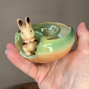 May include: A ceramic bunny figurine in a decorative dish. The bunny is tan with brown accents, and the dish is green and tan. The dish has a unique shape with a small, raised section.