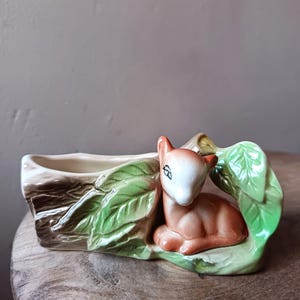 May include: A ceramic planter featuring a fawn nestled among green leaves. The planter is shaped like a log with a cream-colored interior. The fawn is brown and white, adding a touch of nature-inspired decor.