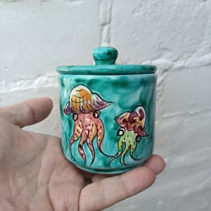 May include: A small, turquoise ceramic jar with a matching lid. The jar is decorated with hand-painted jellyfish in shades of orange, pink, and green. The jar is held in a person's hand.