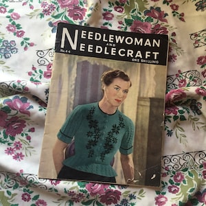 May include: Vintage magazine cover titled "Needlewoman and Needlecraft" featuring a woman in a teal knitted top with black floral embroidery. The magazine cover is slightly worn, with the number 44 visible.