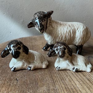 May include: A set of three ceramic sheep figurines featuring one standing adult sheep and two smaller lambs in a resting position. Each figurine is finished in a glossy glaze with cream-colored bodies and dark brown markings on the faces and legs.