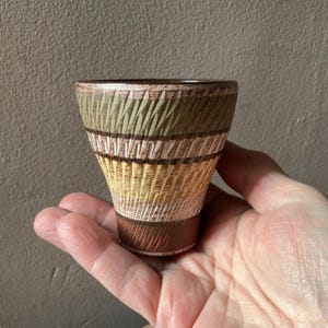 May include: A small, handcrafted ceramic cup with a textured, woven-like design. The cup features bands of colour, including green, brown, yellow, and white. The base is a dark brown, and the rim is a darker shade of brown.