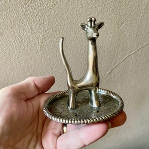 May include: A silver-colored giraffe-shaped ring holder. The giraffe has a long neck and a small head with detailed features. The ring holder has a round base with a floral pattern and a beaded edge. The ring holder is held in a hand.