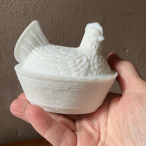 May include: A white milk glass hen on a nest dish. The lid is shaped like a sitting hen with detailed feather textures, while the base features a molded basket weave pattern. This opaque white glass container is a classic lidded decorative piece.