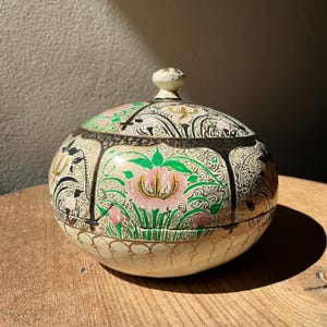 May include: A round, lidded decorative box with a floral design. The box is cream-colored with pink, green, and gold accents. The lid features a knob and intricate patterns. The box is sitting on a wooden surface.
