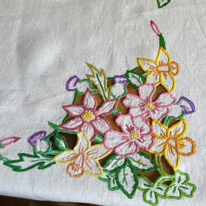 May include: A white linen tablecloth featuring intricate cutwork embroidery in the corner. The design displays a cluster of flowers in shades of pink, yellow, and purple, accented with green leaves and stems, showcasing detailed needlework.