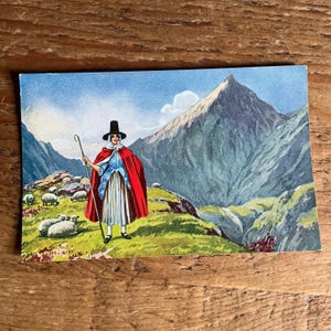 May include: A vintage postcard depicting a shepherdess in traditional dress, a red cloak, and a staff, with sheep grazing in a mountainous setting. The background shows a towering mountain range under a blue sky.