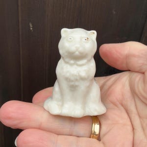 May include: A small, white ceramic cat figurine with a sitting pose. The cat has a detailed face and a textured chest. The figurine is held in a hand, showcasing its size and design.