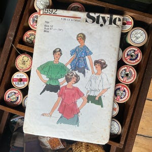 May include: A vintage "Style" sewing pattern with illustrations of women's tops. The pattern includes size and measurement details, with a bust measurement of 87 cm. Wooden spools of thread in various colors surround the pattern.