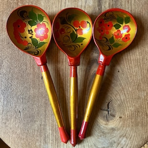 May include: Three wooden spoons with a traditional Russian Khokhloma design. The spoons have a gold bowl with red floral patterns and green leaves. The handles are gold with red tips.