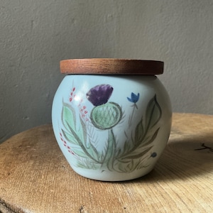 May include: A round, light blue ceramic jar with a smooth finish and a flat, circular wooden lid. The exterior features a hand-painted illustration of a green thistle with a purple flower head, surrounded by green leaves and small red berries.