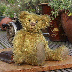 May include: A golden-brown teddy bear with shaggy fur, dark eyes, and a brown stitched nose. The bear has light brown felt-like fabric on the soles of its feet and sits in an upright position with its legs extended forward on a wooden surface.