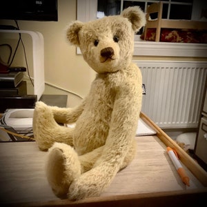 Può includere: Un grande orsacchiotto di peluche beige con naso marrone e occhi neri è seduto su una superficie di legno. L'orso ha una consistenza soffice e usurata ed è seduto con le gambe incrociate.