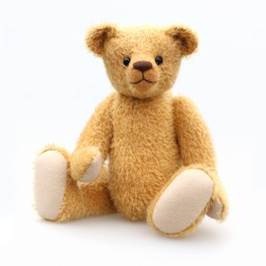 May include: A plush, light brown teddy bear with a soft, fuzzy texture. The bear has a brown nose and black eyes. It is sitting with its legs crossed and its paws resting on the ground.