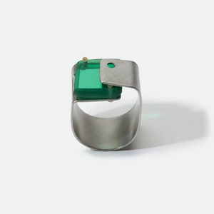 Könnte beinhalten: Moderner Ring mit einem gebürsteten Silberband und einem quadratischen, smaragdgrünen Edelstein. Der Edelstein ist in einen silbernen Rahmen gefasst, akzentuiert durch ein kleines goldfarbenes Detail. Der Ring hat ein modernes, minimalistisches Design.