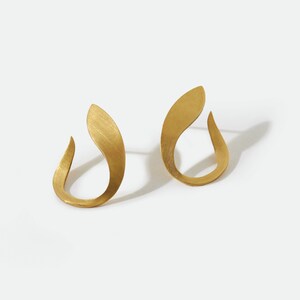 May include: A pair of gold-colored earrings with a curved, abstract design. Each earring has a smooth, matte finish and a teardrop shape, with a hook at the bottom. The earrings are displayed on a white background.