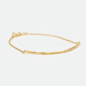Puede incluir: Una pulsera dorada con un diseño de barra texturizada. La pulsera presenta una delicada cadena y una pieza central en forma de barra con una superficie irregular. La joya está sobre un fondo blanco.