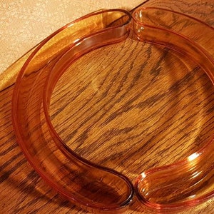 May include: A transparent, amber-coloured, curved glass object, possibly a decorative bowl or serving piece. The piece is resting on a wooden surface, with a patterned fabric in the background. The glass has a smooth, glossy finish.