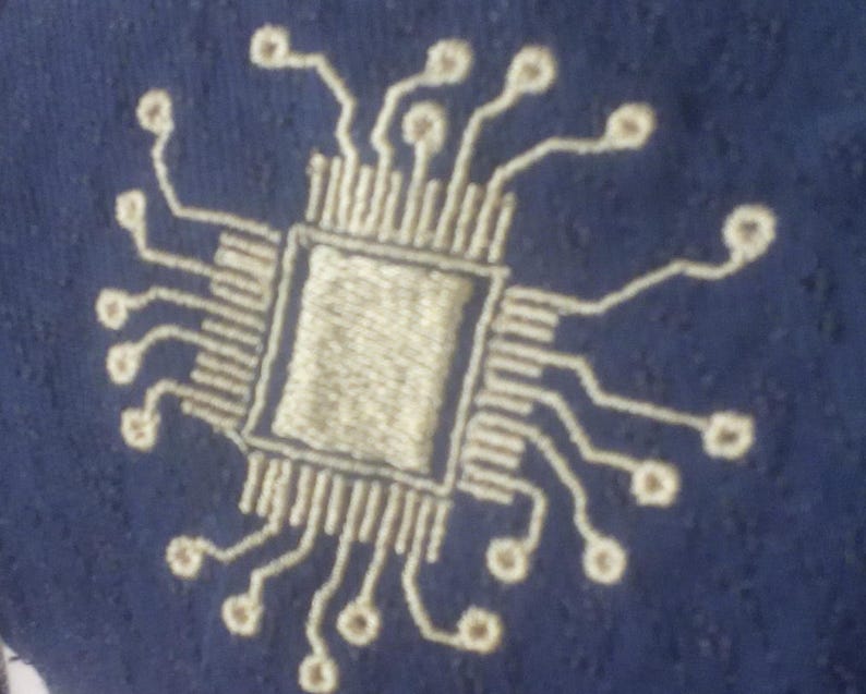 May include: A white embroidered design of a computer chip on a dark blue fabric background.