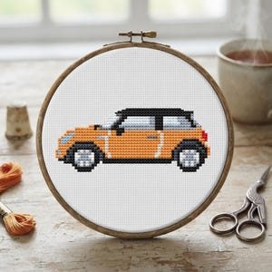 May include: A cross-stitch embroidery project featuring a side profile of an orange compact car with a black roof and silver wheels, displayed within a circular wooden hoop on a white fabric background, showcasing detailed pixelated stitching.